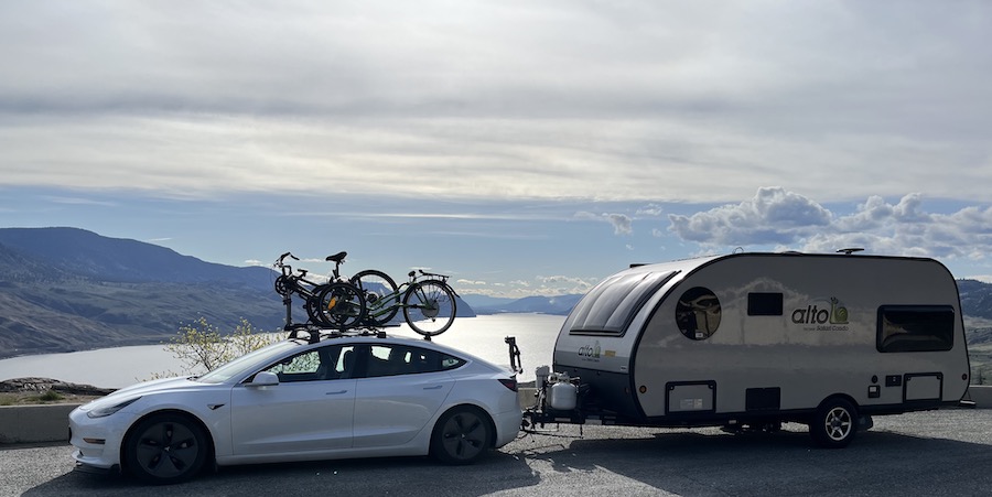 Tesla model 3 with bicycle rack pulling travel trailer on road in front of lake and mountains